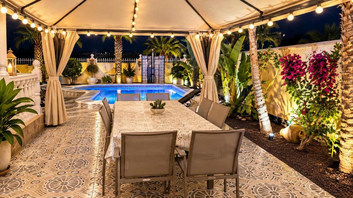 An illuminated outdoor dining area with a table and chairs overlooks a swimming pool surrounded by palm trees and tropical plants.
