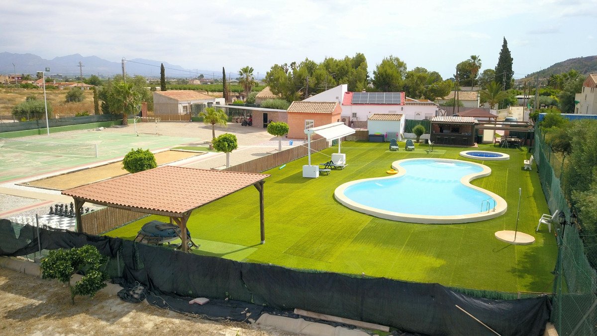 Aerial view of a villa with a large freeform swimming pool, a smaller jacuzzi, a tennis court, and outdoor seating areas.