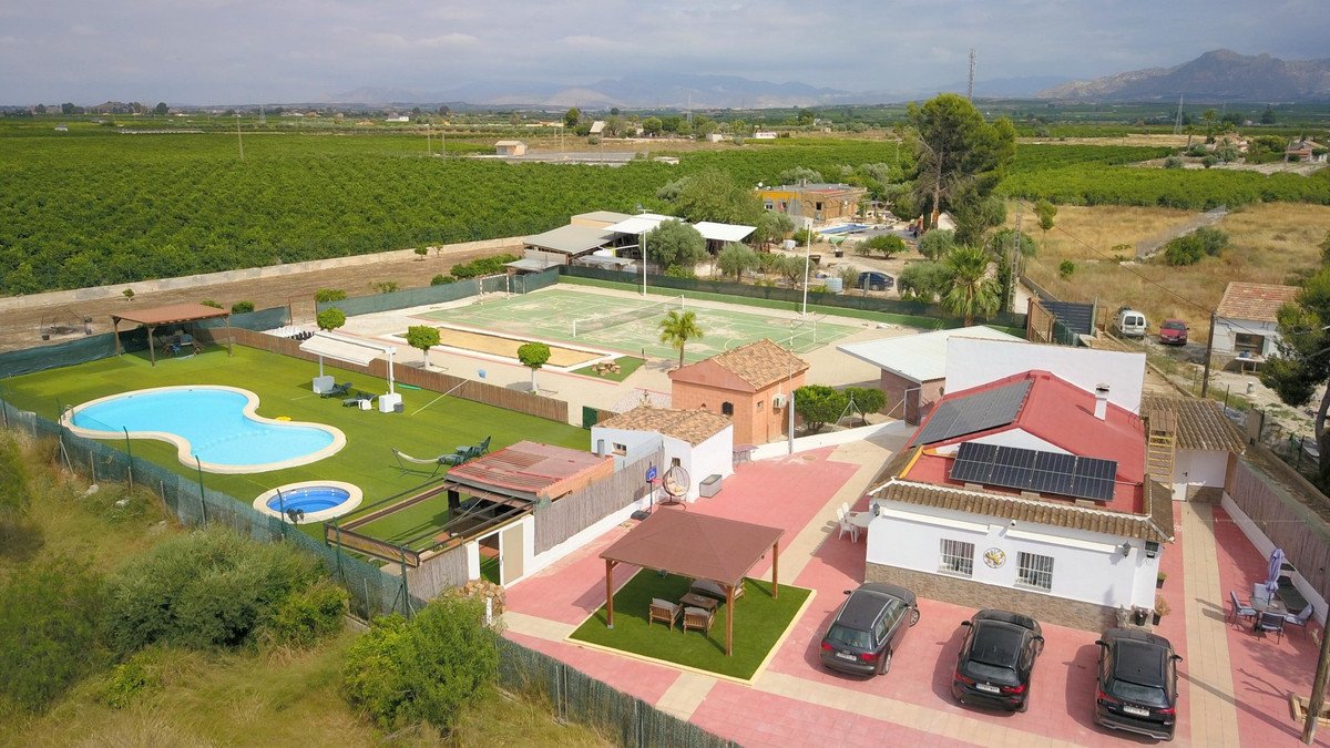Aerial view of a property featuring a swimming pool, a tennis court, and surrounding agricultural fields under a cloudy sky.