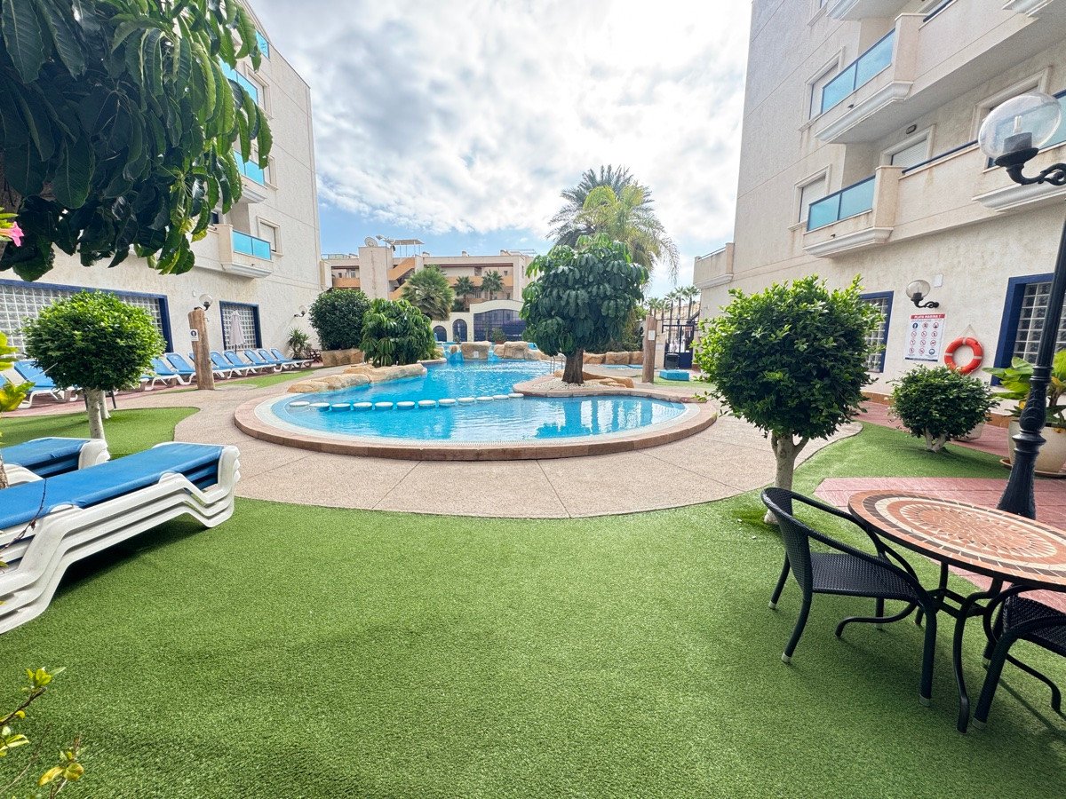 Outdoor communal pool area with artificial turf, sun loungers, palm trees, and a patio table with chairs.