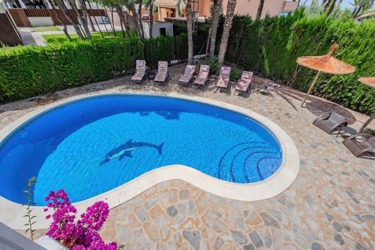 A bright blue kidney-shaped swimming pool with a dolphin mosaic at one end, surrounded by a stone patio and green hedges. Sunbeds are visible.