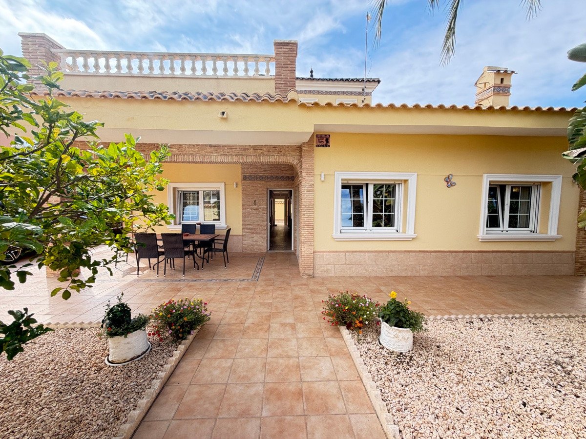Yellow villa with a tiled terrace, outdoor dining area, and garden with plants and trees. Blue sky above.