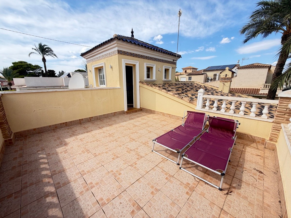 Terrace with two purple lounge chairs, a small building with a blue tiled roof, and palm trees under a blue sky.
