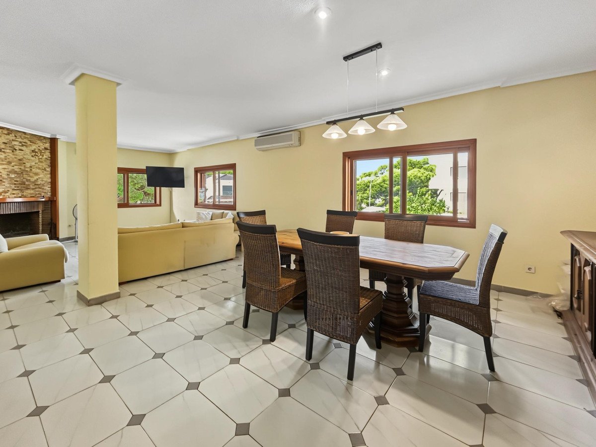 Spacious living and dining room with a fireplace, comfortable sofa, dining table, and large windows offering a green view.