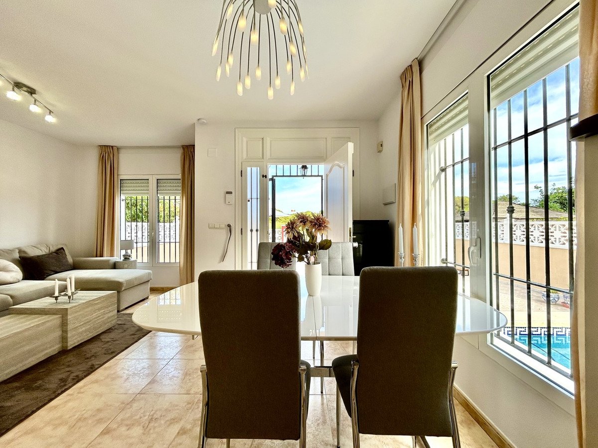 Modern dining and living area with a sofa, dining table, and chairs, overlooking a pool through large windows.