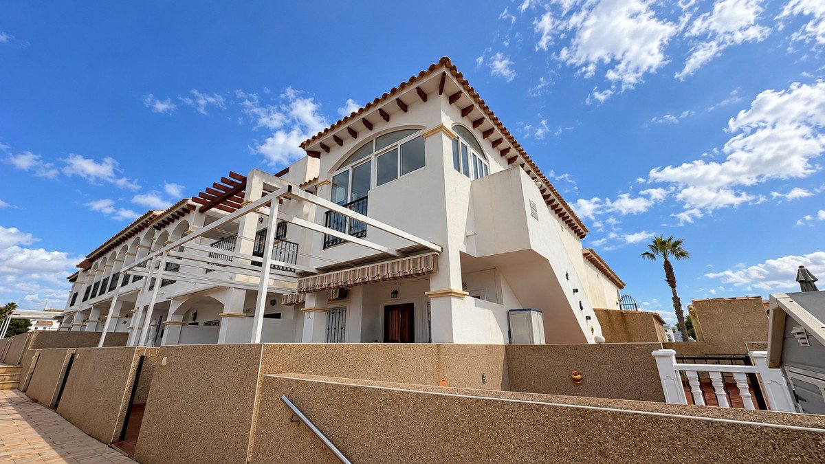 Exterior view of a multi-story villa with arched windows, balconies, awnings, and a palm tree under a blue sky with clouds.