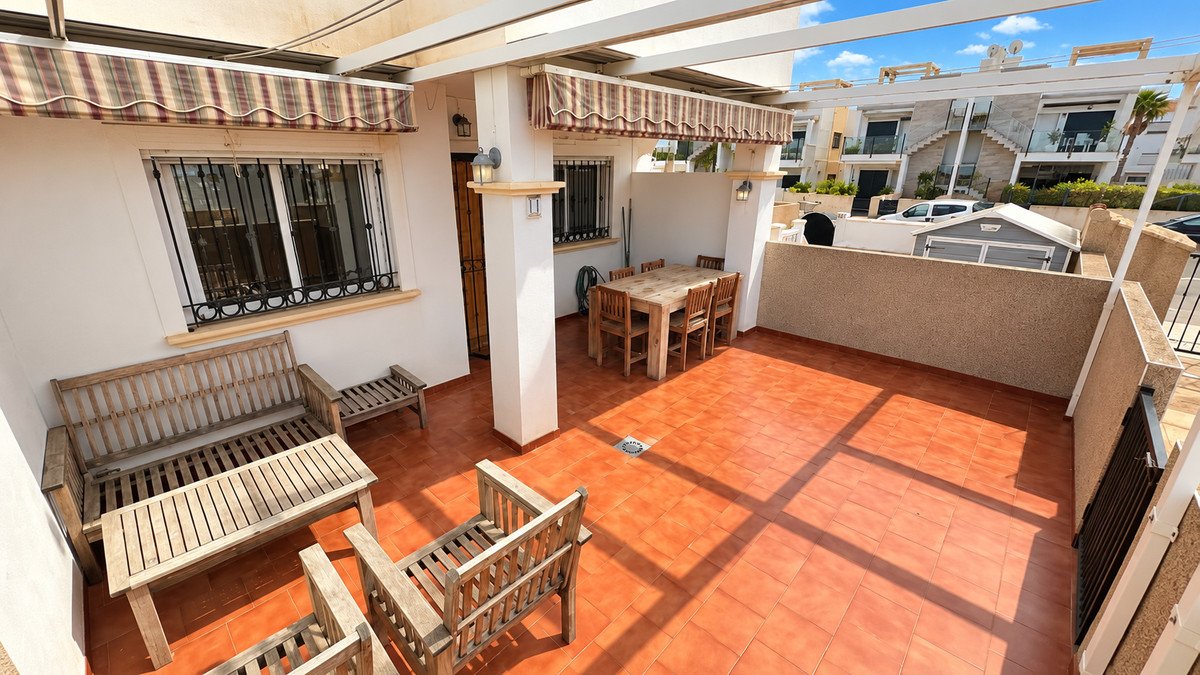 Sunny terrace with wooden dining set and lounge seating, featuring striped awnings and a view of houses.