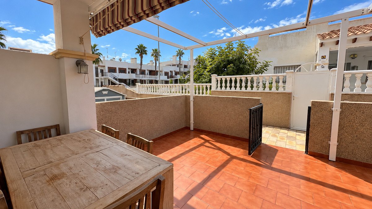 A sunny terrace with a wooden dining set, featuring palm trees and buildings in the background under a clear blue sky.
