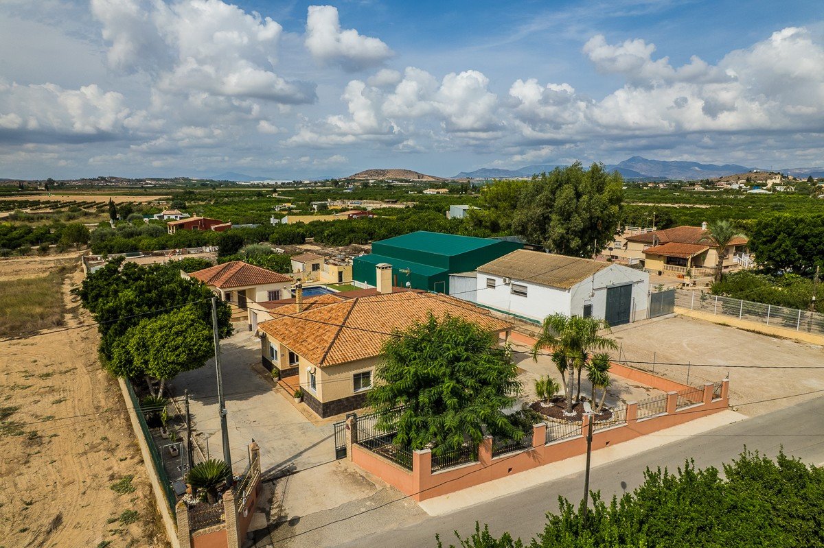 Aerial view of a rural property with a villa, swimming pool, and surrounding landscape under a cloudy sky.