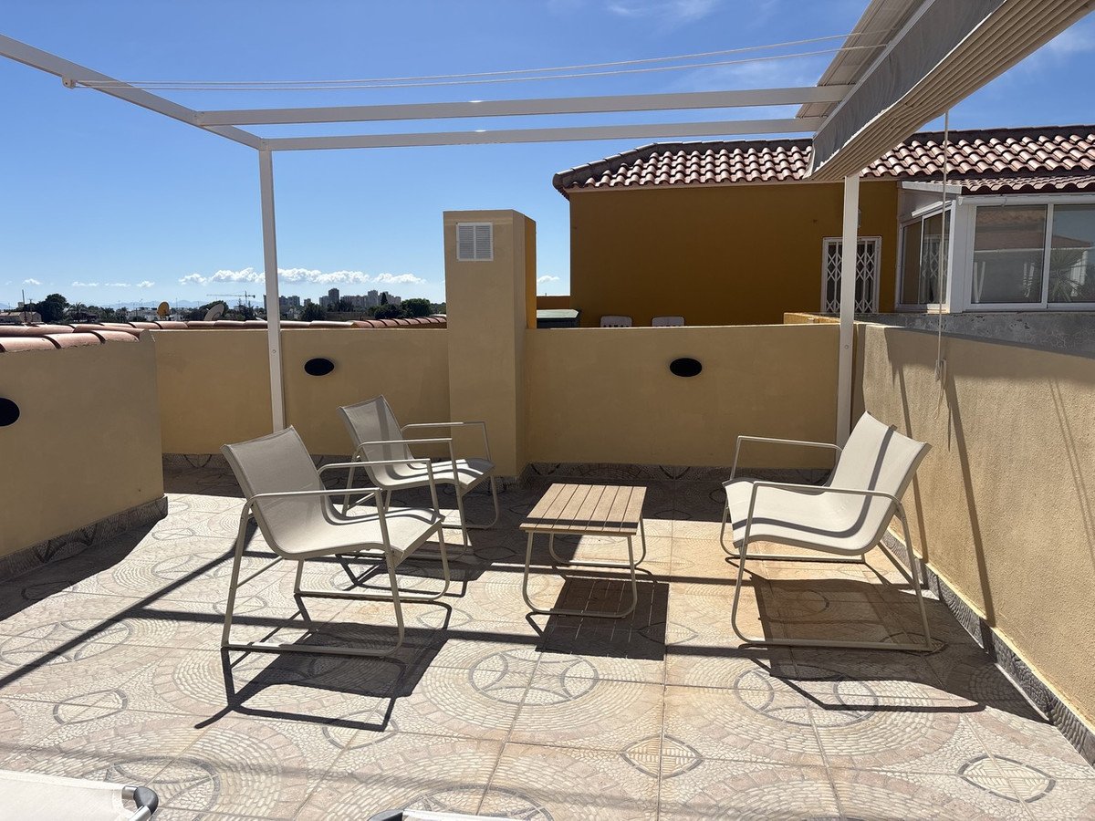 Terrace with chairs and a table overlooking a cityscape on a sunny day. White pergola structure overhead.