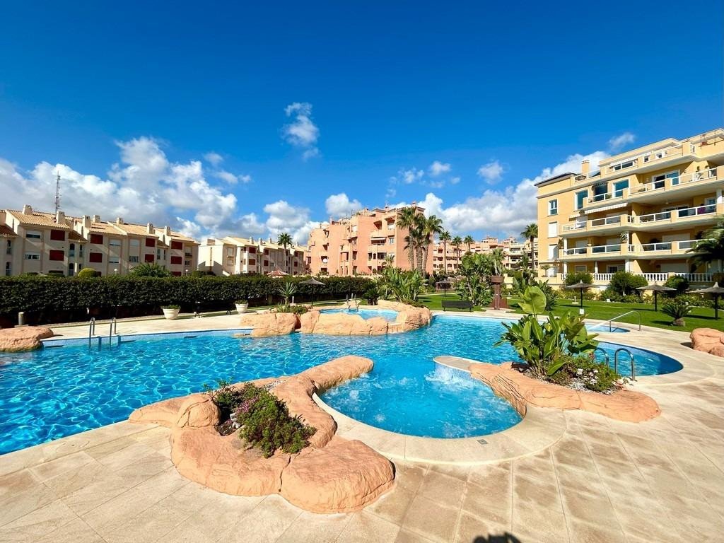 A bright outdoor swimming pool and jacuzzi area with sun loungers and apartment buildings in the background under a blue sky.