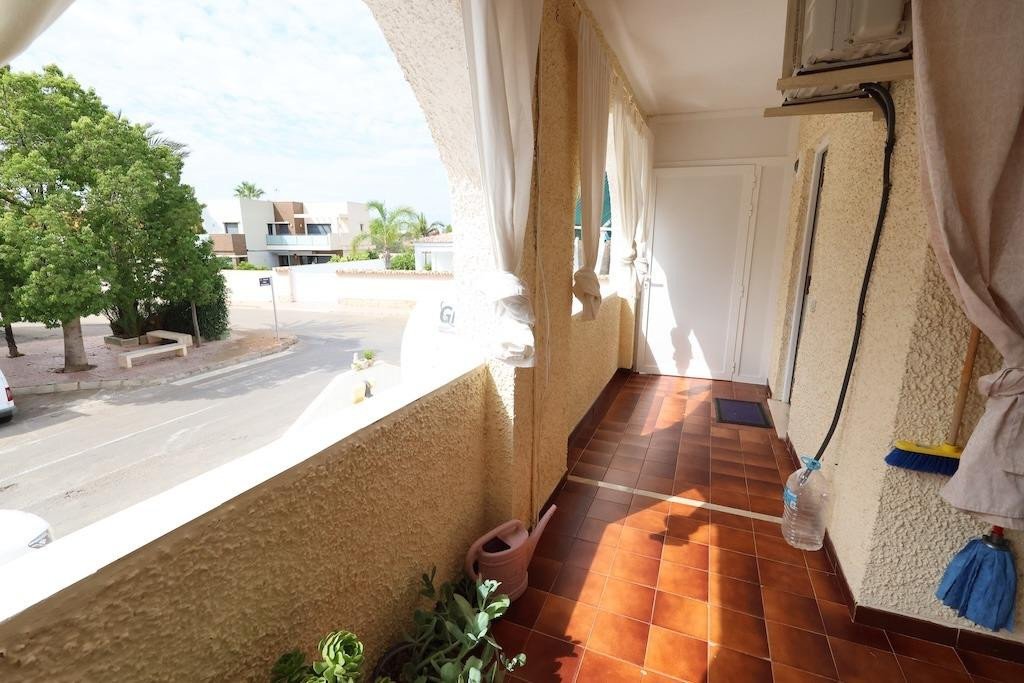 Sunny balcony overlooking a street with trees and modern houses, featuring terracotta tiles and white curtains.