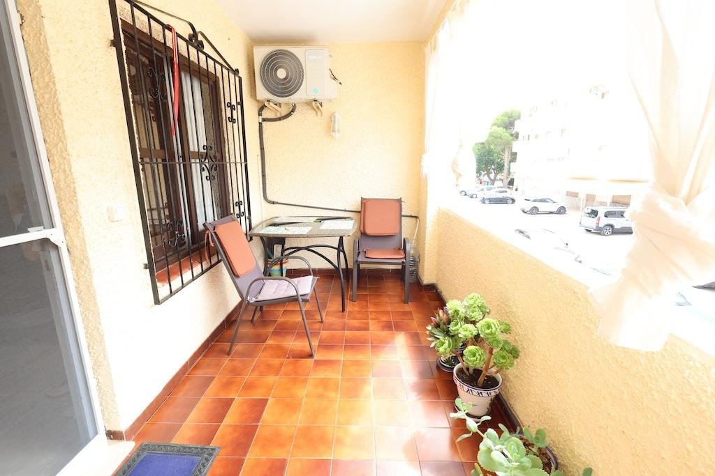 A balcony with two chairs, a table, an air conditioner, and potted plants, overlooking a street with cars.