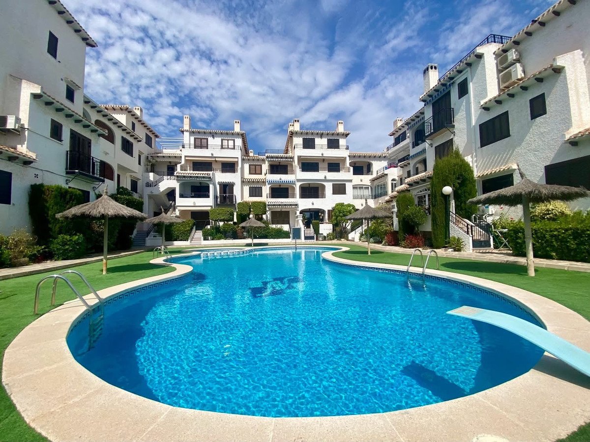 A sparkling blue swimming pool with a diving board surrounded by green artificial grass and apartment buildings under a cloudy blue sky.