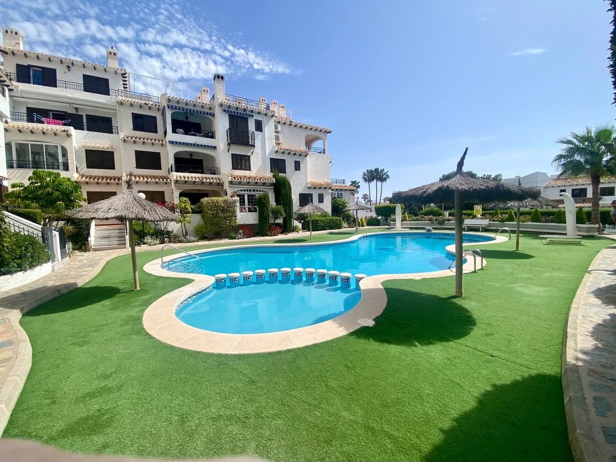 Exterior view of an apartment complex with a swimming pool, sun loungers, and umbrellas on a sunny day.
