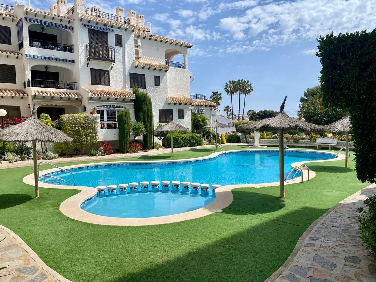 Resort complex with a large swimming pool, sun loungers, parasols, and lush green surroundings on a sunny day.