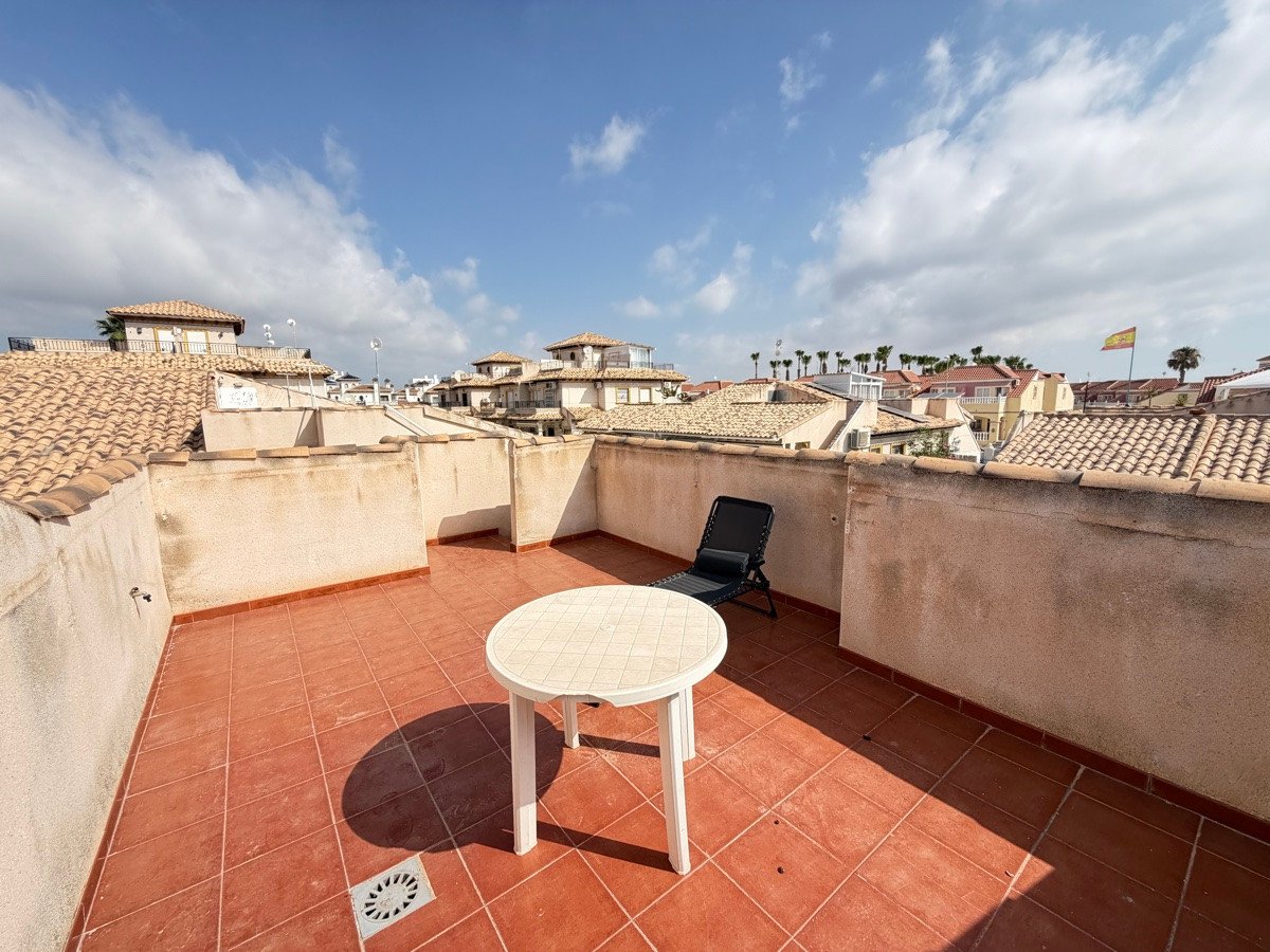 Terrace with red tiled floor, outdoor table, chair, and a town view under a blue sky with clouds.