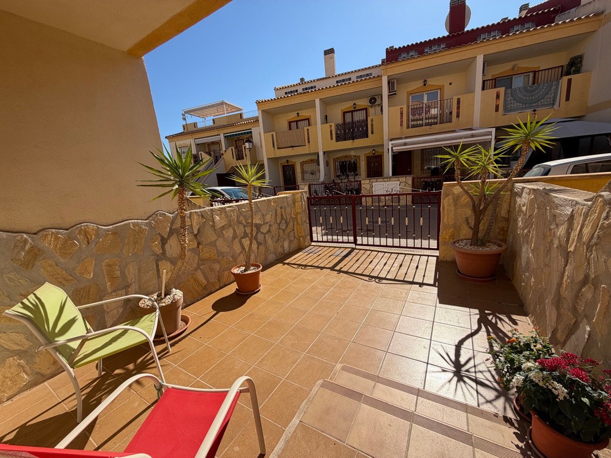 A sunny terrace with two lounge chairs, potted plants, and a view of attached houses and a gate.