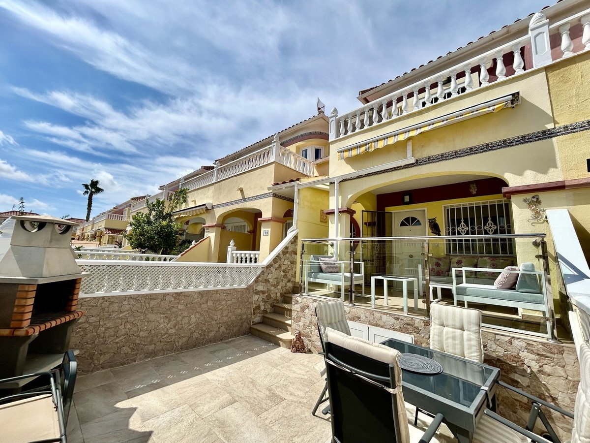 A sunny terrace with an outdoor kitchen, dining set, and seating area in front of a yellow townhouse with balconies.