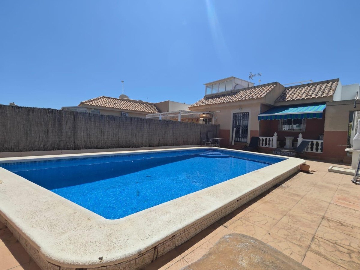 Backyard pool with tiled deck, patio furniture, and house under clear blue sky.