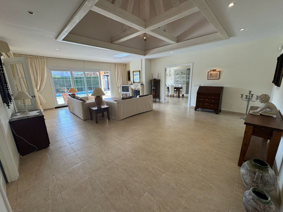 Spacious living room with high beamed ceiling, tiled floors, and large windows overlooking a pool.