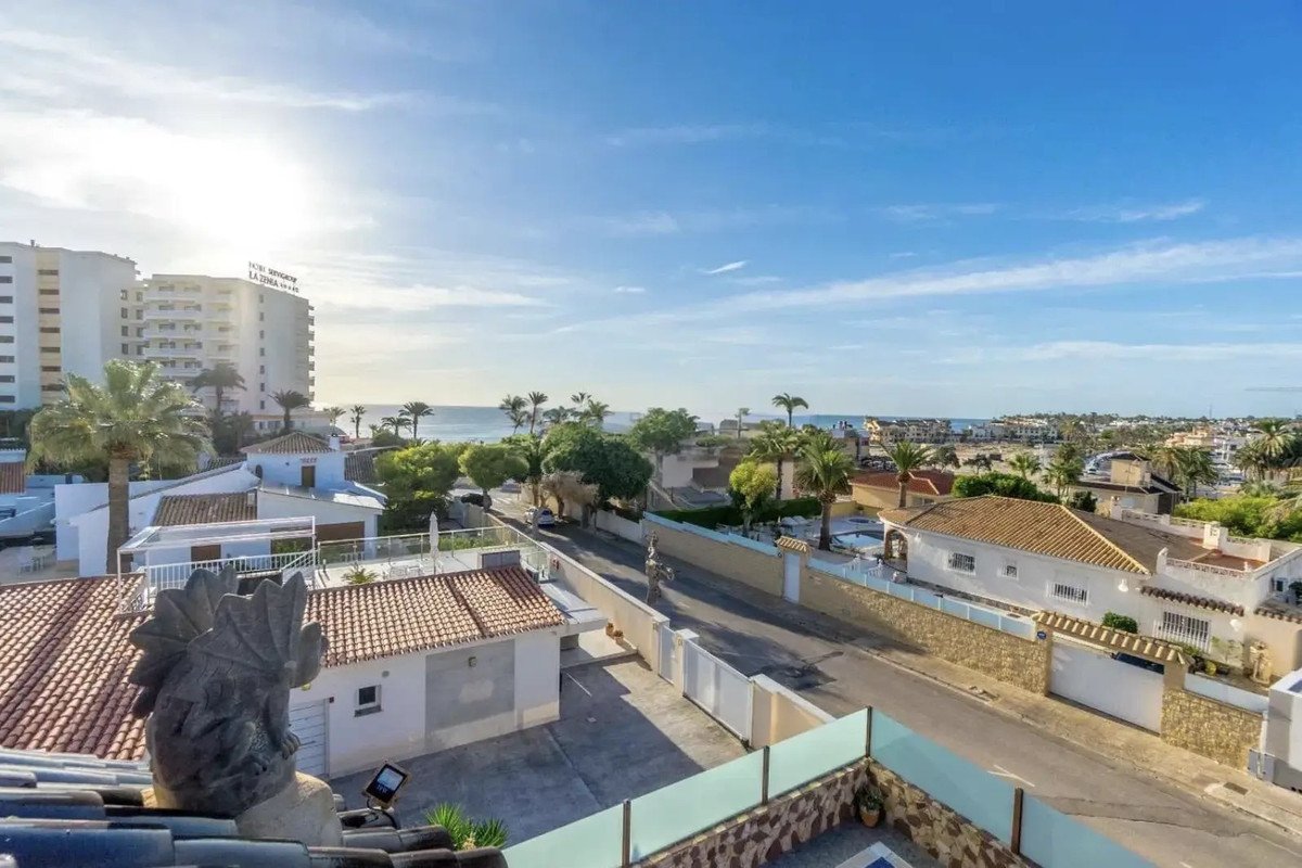 View from a balcony overlooking rooftops, a street, palm trees, and the sea in the distance.