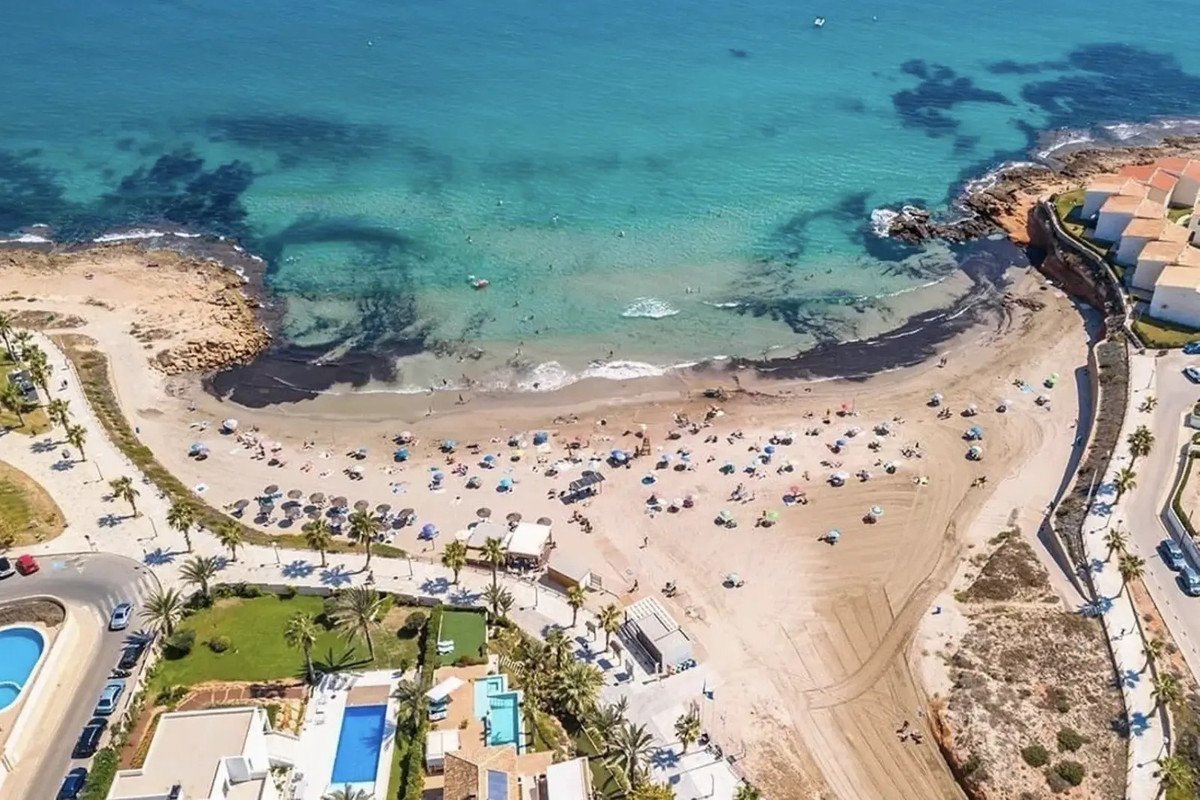 Aerial view of a crowded beach with turquoise water, palm trees, and modern buildings with pools.