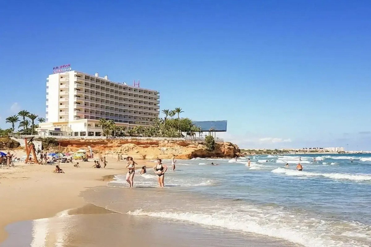 A large hotel building overlooks a sandy beach with people swimming and relaxing by the ocean on a sunny day.