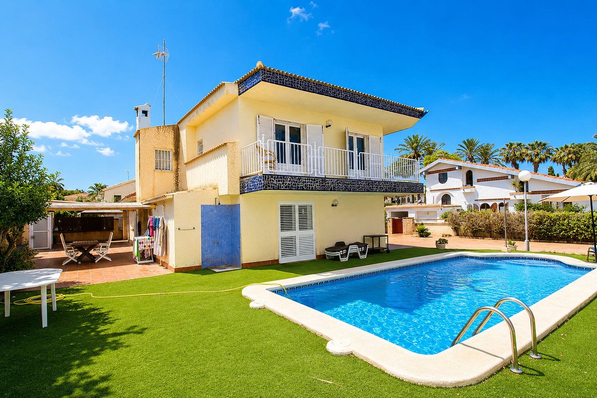 Yellow villa with a swimming pool, artificial grass lawn, and a tiled patio area with outdoor dining furniture.