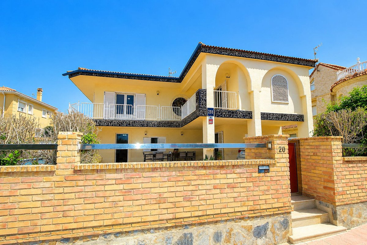 A yellow villa with white balconies and arched windows, viewed from behind a brick fence on a sunny day.
