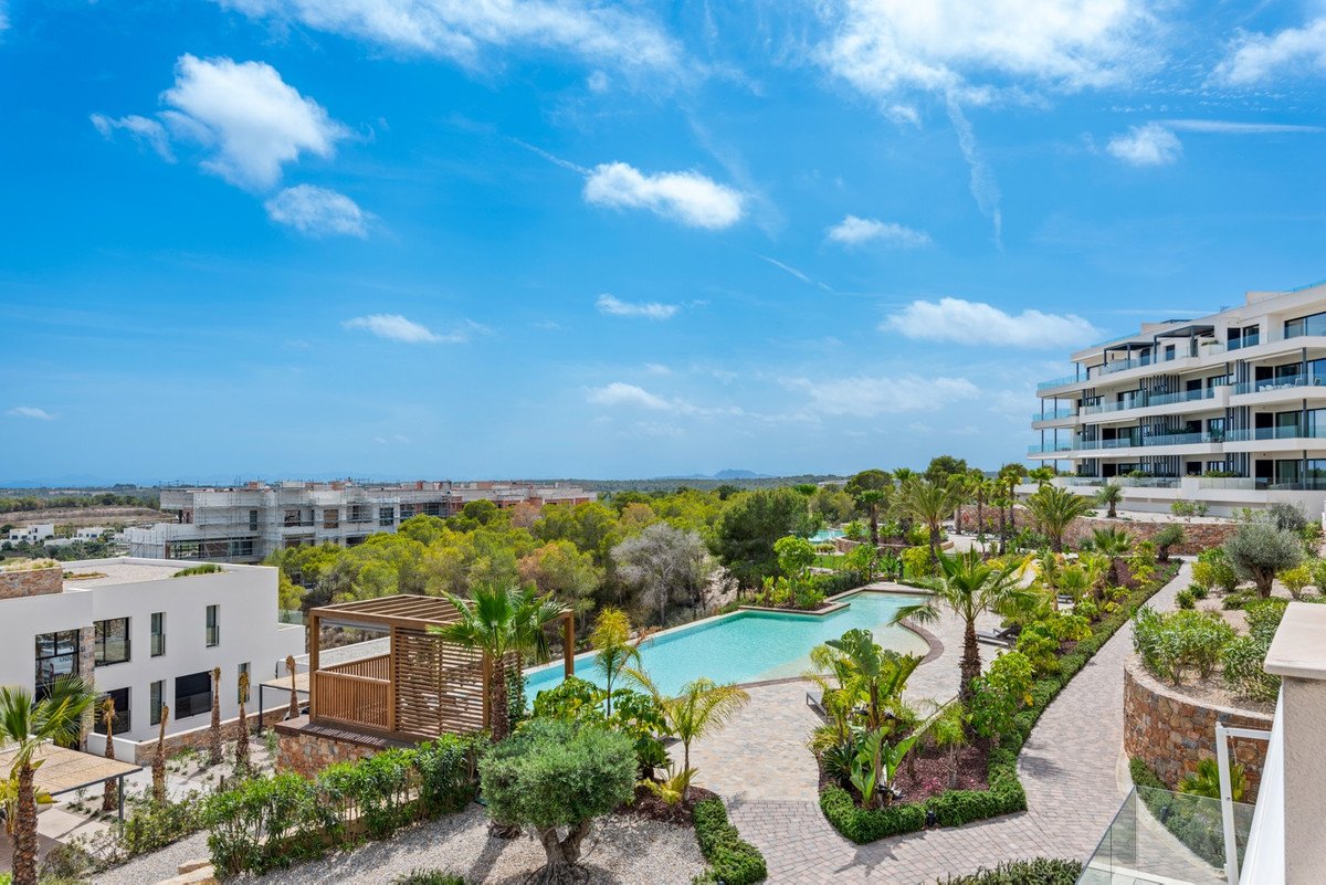 A resort complex featuring a curved swimming pool surrounded by lush greenery, palm trees, and modern apartments with balconies.