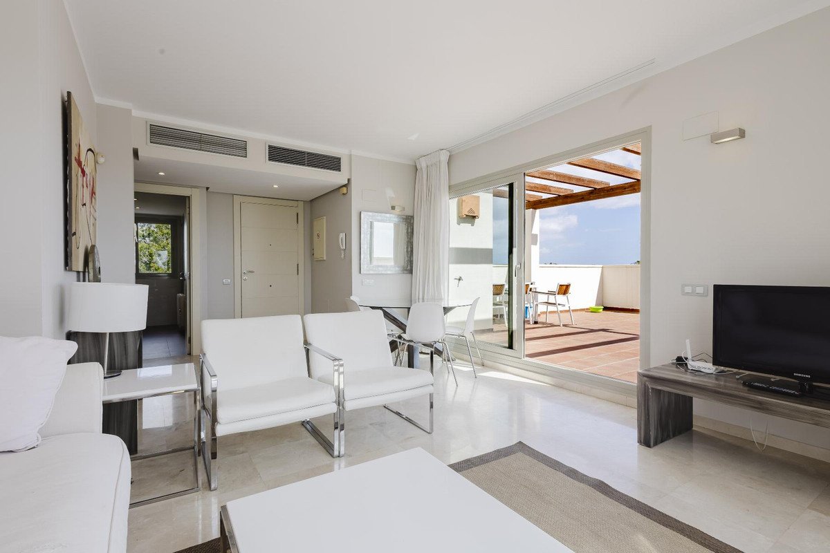 Bright living room with modern white furniture, large sliding doors opening to a sunny terrace with outdoor seating.