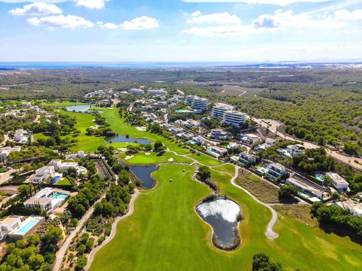 Aerial view of luxury villas and modern buildings situated on a lush golf course with lakes, overlooking the ocean.