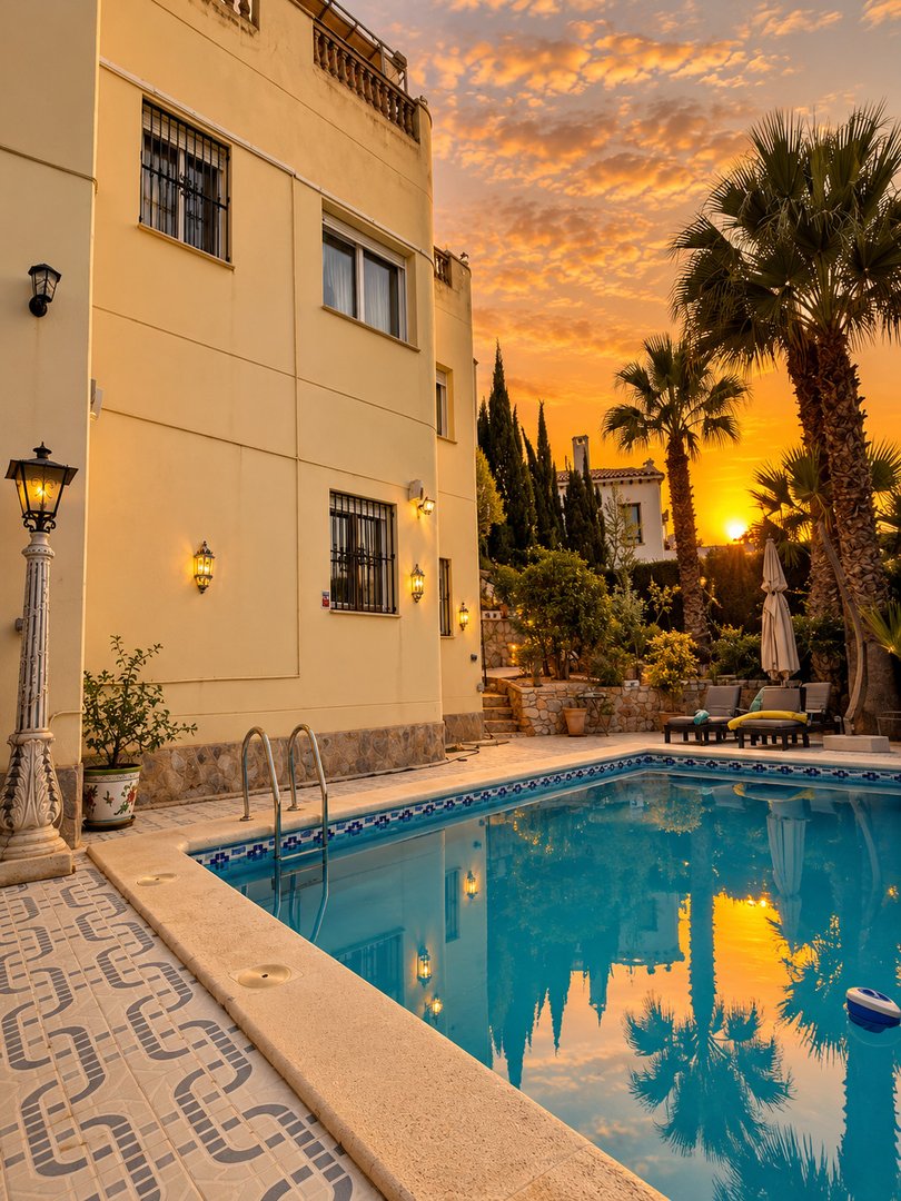 A villa with a pool and palm trees during a vibrant sunset, reflecting in the water.