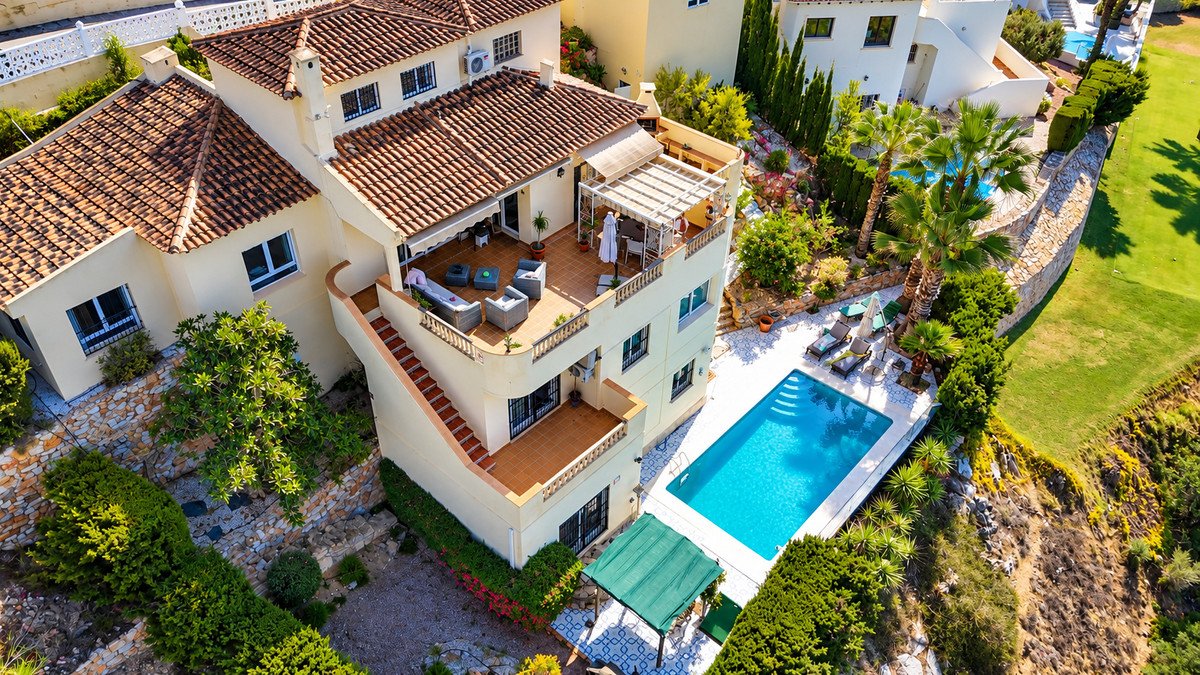 Aerial view of a multi-story villa with a swimming pool, sun loungers, multiple terraces, and lush greenery, bordering a golf course.