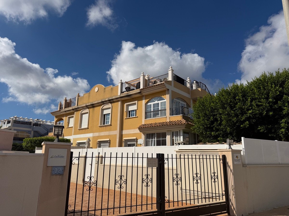 Exterior of a yellow villa with arched windows, balconies, a terrace, and a front gate under a blue sky with clouds.