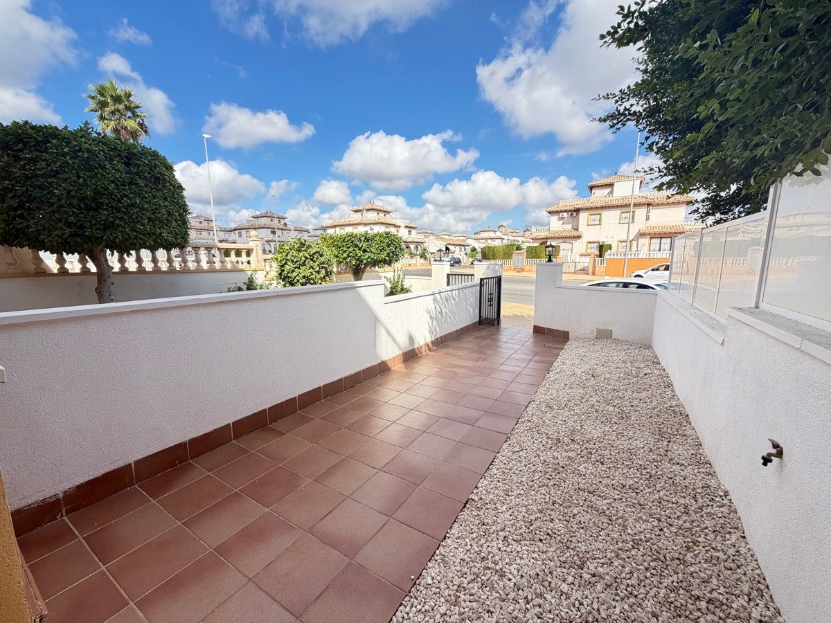 A tiled terrace and gravel area outside a property, with a white wall, gate, and houses in the background under a blue sky.