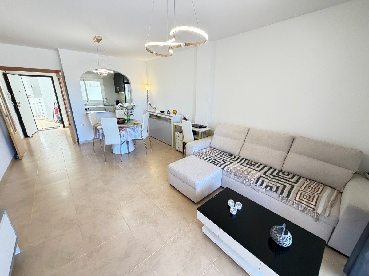 Modern living room with a sofa, coffee table, dining area, and open-plan kitchen. Features bright lighting and contemporary decor.