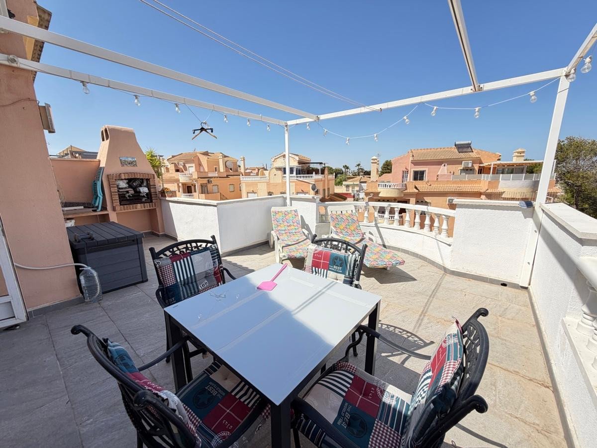 Rooftop terrace with dining table, chairs, barbecue, lounge chairs, and a view of residential buildings.
