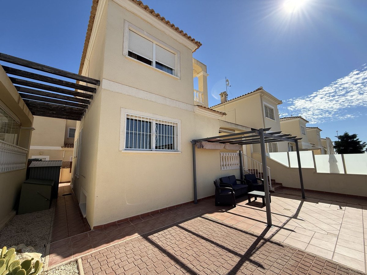 A sunny terrace with seating area under a pergola, next to a house with white shutters and security bars.