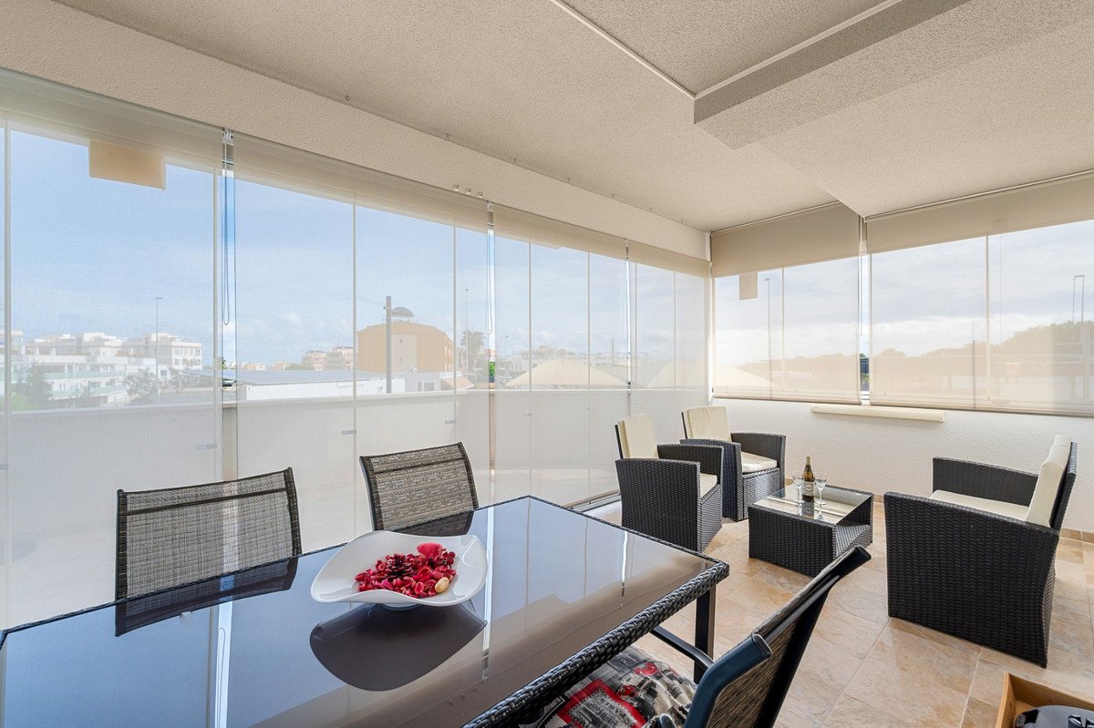 Balcony dining area with glass walls and sea view, featuring wicker furniture and a glass table.