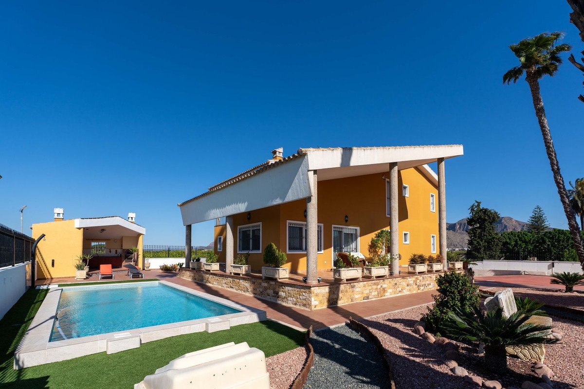 Yellow villa with a swimming pool, covered patio, outdoor kitchen area, and mountain views under a clear blue sky.