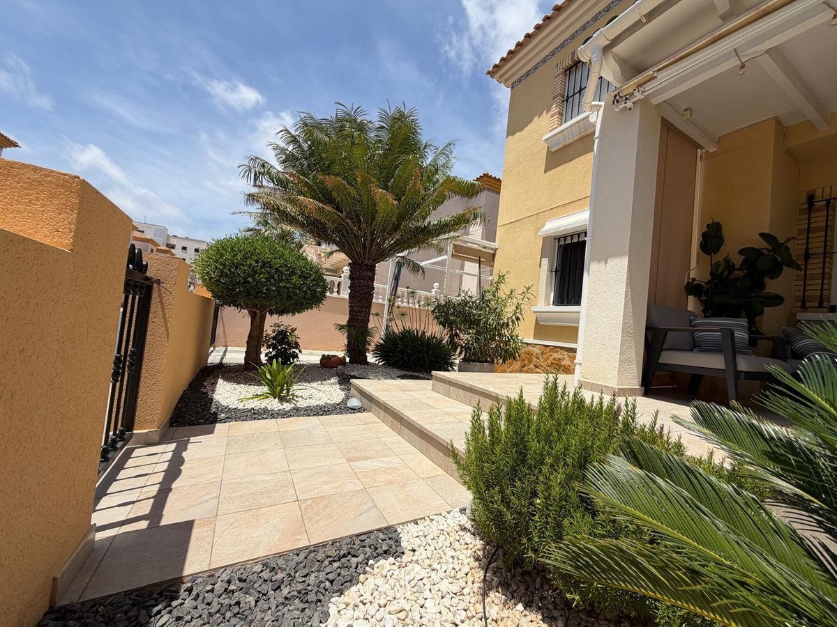 A paved pathway leads to a house entrance, surrounded by a garden with a palm tree, gravel, and lush greenery.