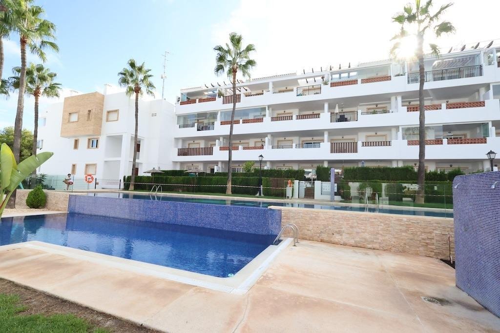 A modern apartment building with balconies overlooks a large swimming pool surrounded by palm trees and lush greenery.