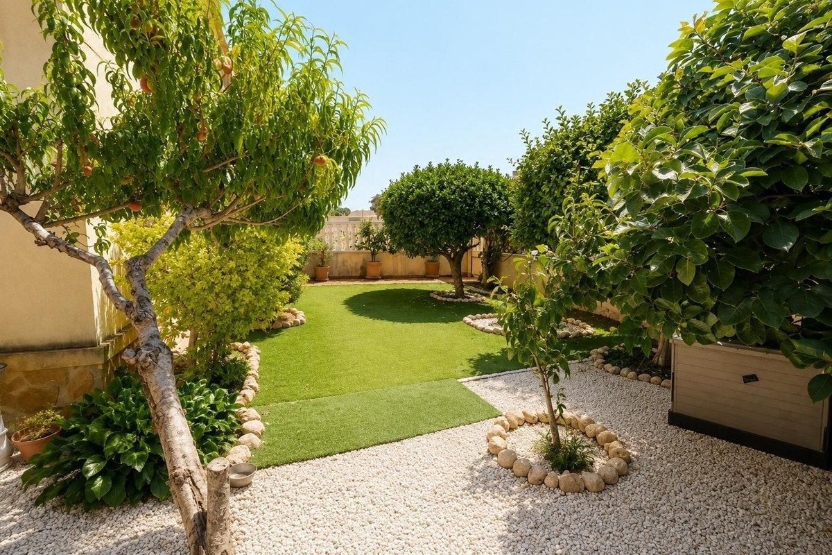 A beautifully landscaped garden with a green lawn, mature trees, and white pebble pathways on a sunny day.