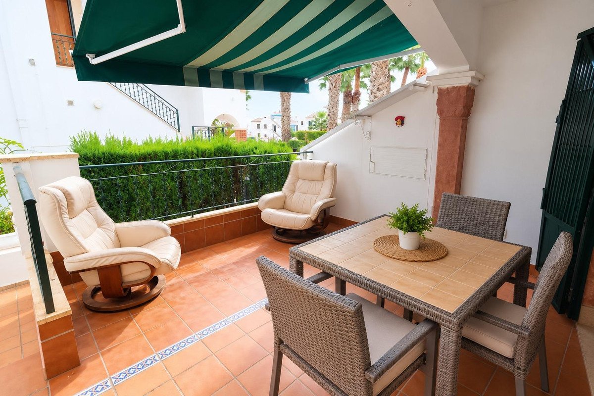 A covered terrace with outdoor dining table, chairs, and comfortable seating, offering a view of greenery and buildings.