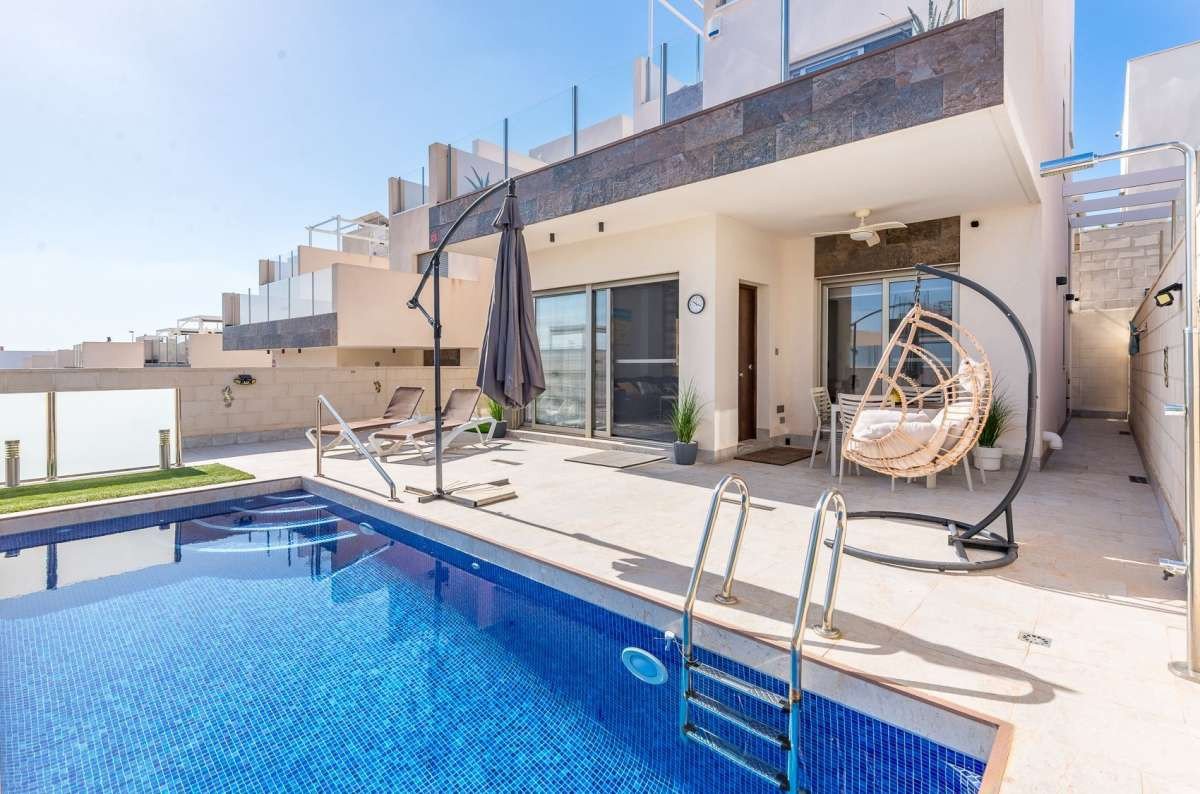 Private swimming pool and patio with sun loungers and a hanging chair, leading to a modern villa.