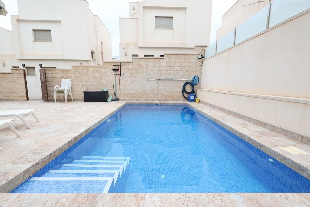 Exterior shot of a private swimming pool with tiled steps, surrounded by a tiled deck and modern architecture. Includes loungers and pool cleaning equipment.