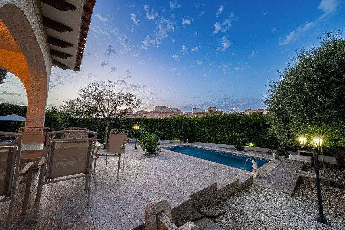 An outdoor patio area with a swimming pool, dining furniture, and garden lights at sunset under a blue sky with clouds.