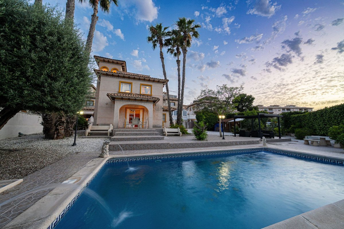 A villa with a swimming pool and patio, surrounded by palm trees, under a cloudy sky at dusk.
